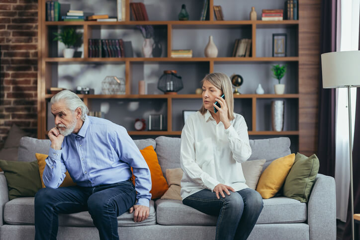 senior married couple, quarreling, gray-haired man and woman sitting on sofa, wife annoyed talking on the phone