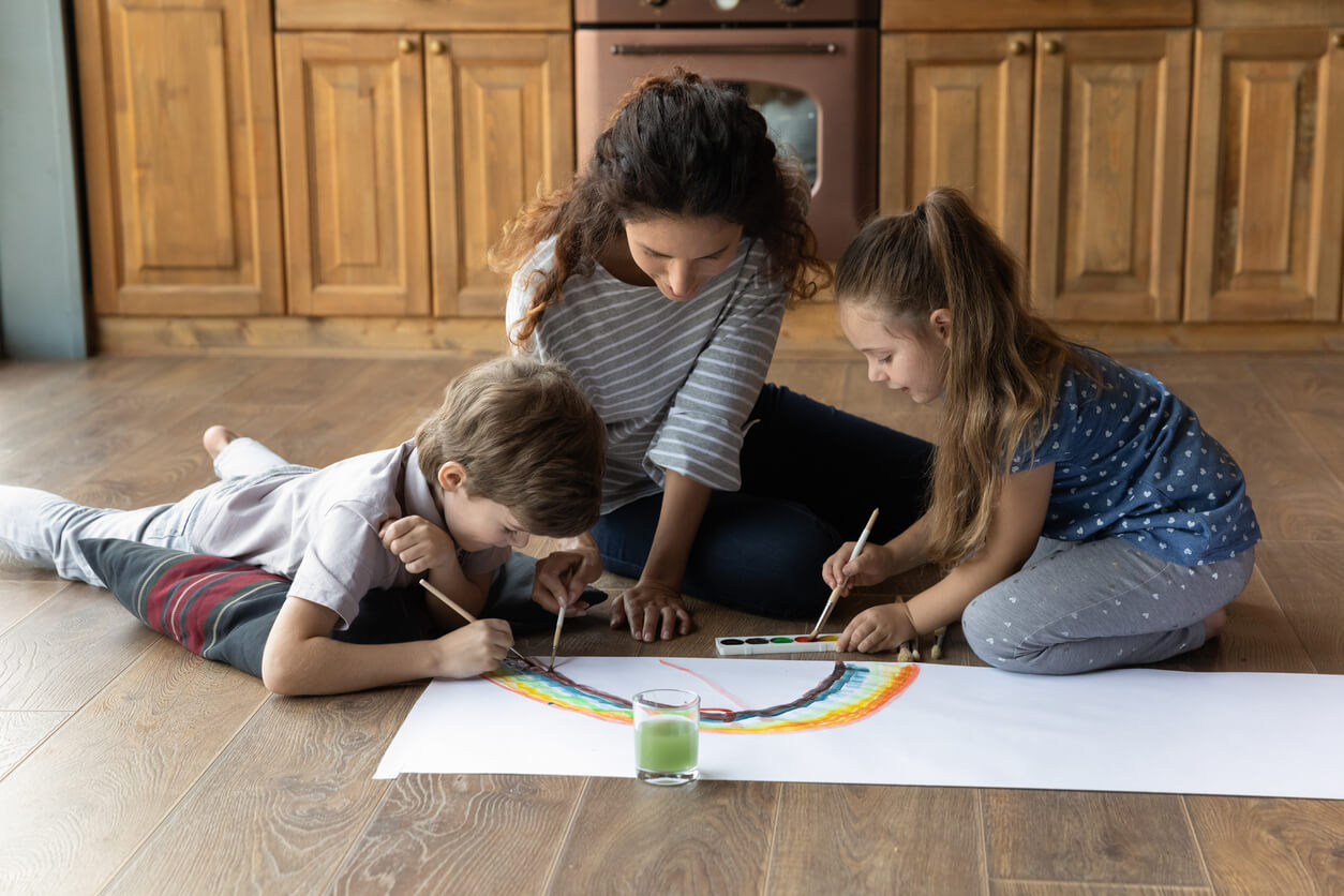 Loving young mother and small children sit on floor in kitchen have fun painting together. Caring happy mom and little preschooler kids engaged in creative funny activity drawing with colors at home.