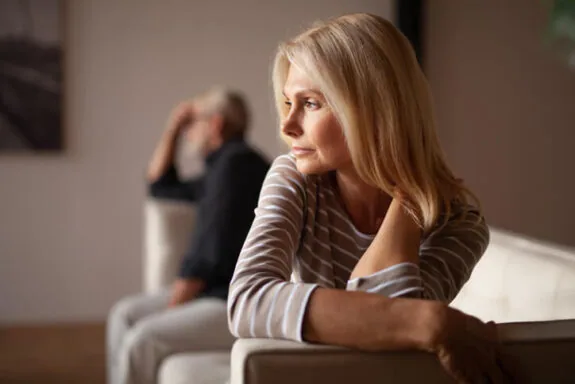 A blonde woman with a pensive expression looks away from a man sitting on a sofa behind her.