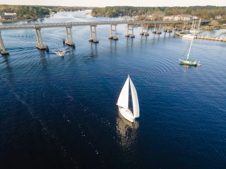 An aerial view of a sailboat on a body of water with a bridge and a marina in the background.